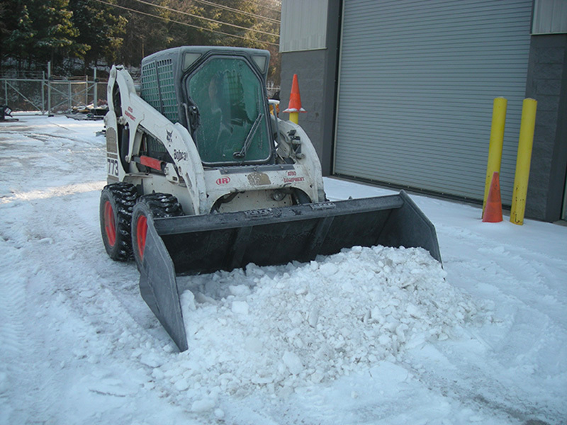left angled view of a skid parked in snow with a blue diamond snow and mulch bucket attachment on the front scooping