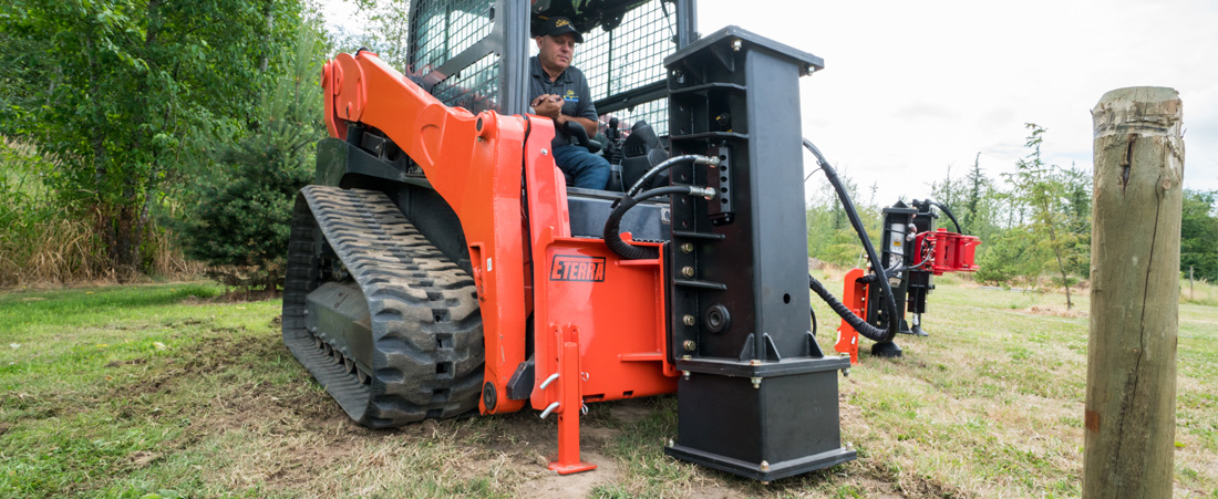 left angled view of a skid steer parked on grass with an eterra skid steer pdx post driver attachment mounted on the front