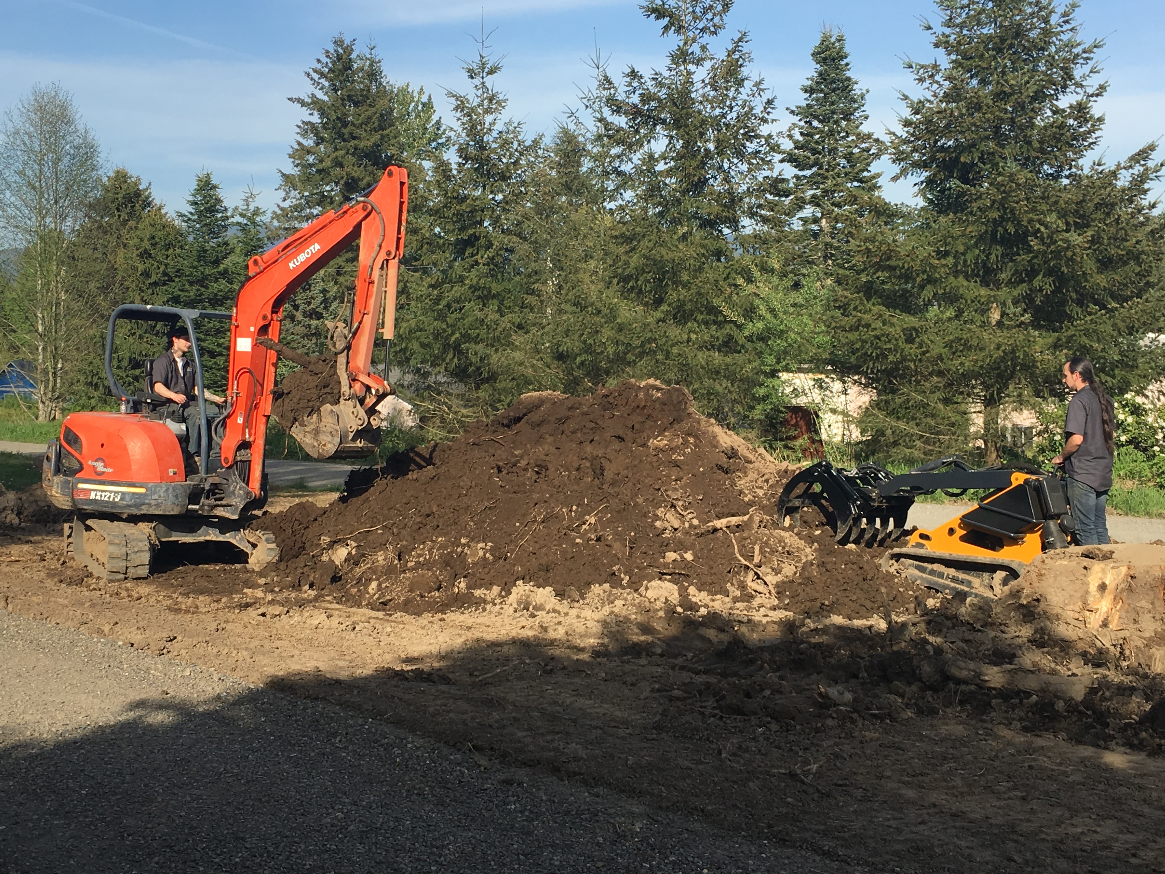 wide view of a skid steer and a mini skid steer parked in a gravel pit across from each with a mound of dirt separating them