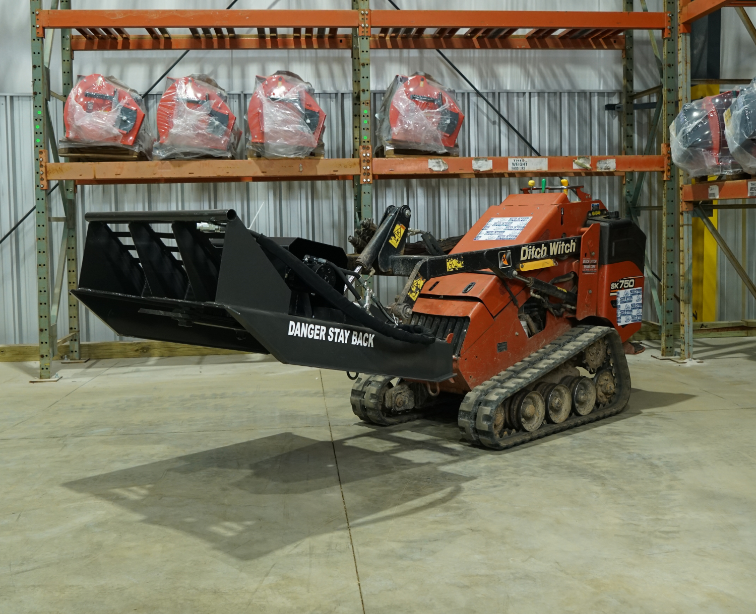 right angled view of a mini skid steer parked in a warehouse with a CID mini skid steer brush cutter attachment on the front raised