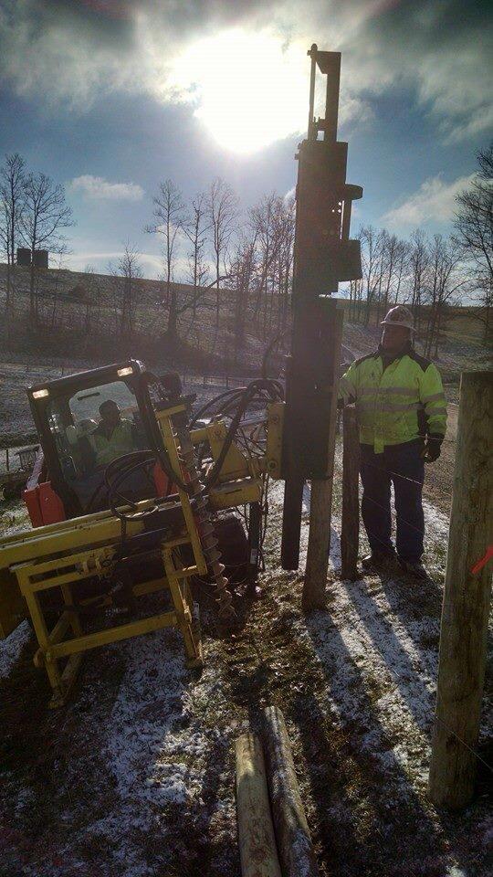 front view of a skid steer parked on angled sloped with a man in safety gear standing to it watching as the operator uses a skid steer post driver to pound a fence post