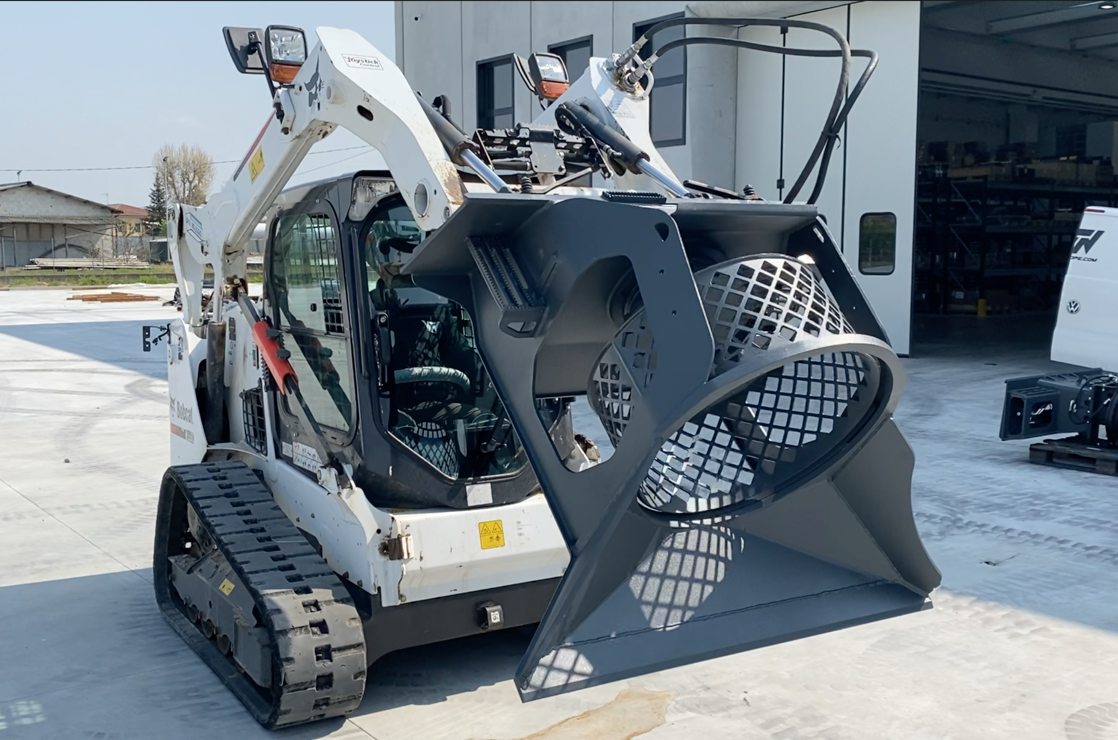 left angled view of a skid steer parked on concrete with a VTN screening bucket attachment on the front lifted up